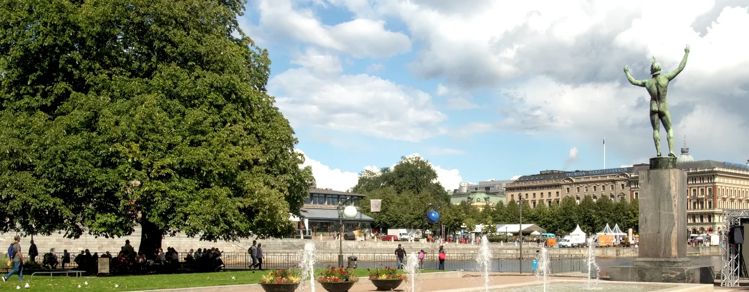 The statue Solsångaren surrounded by summer greenery in Strömparterren. View toward Kungsträdgården, and a glimpse of the Royal Opera in the background. Photo: Rhododendrites (CC BY-SA 4.0)
